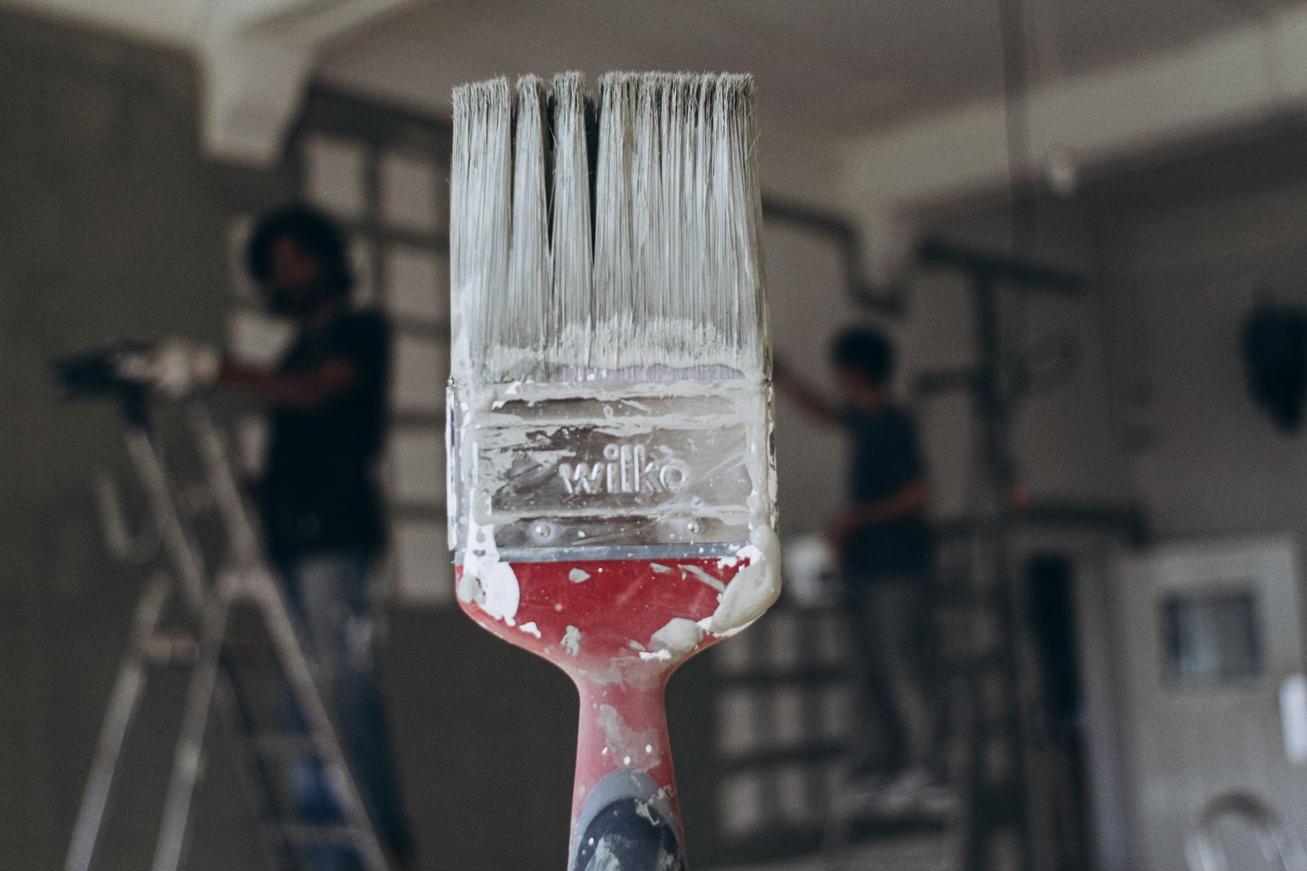 Detailed close-up of a paintbrush with men painting a wall in the blurred background, highlighting renovation work. — Creativ